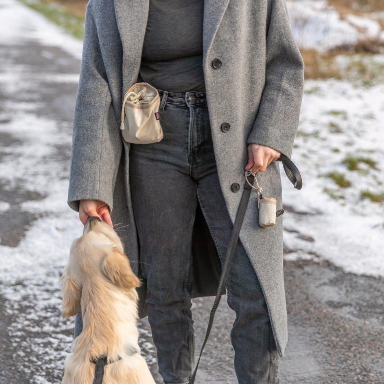 Kvinne står vendt mot kamera med godbitepose til hund synlig på hoften. Hun gir en hund en godbit fra godbitepose til hund fra Trixie.